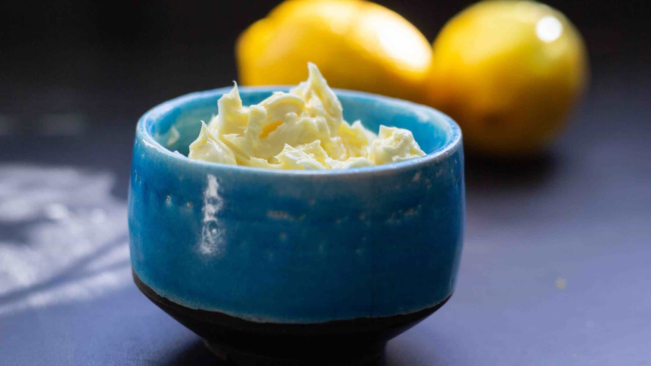 Side view of whipped lemon butter in a turquoise ceramic bowl with two fresh lemons in the background